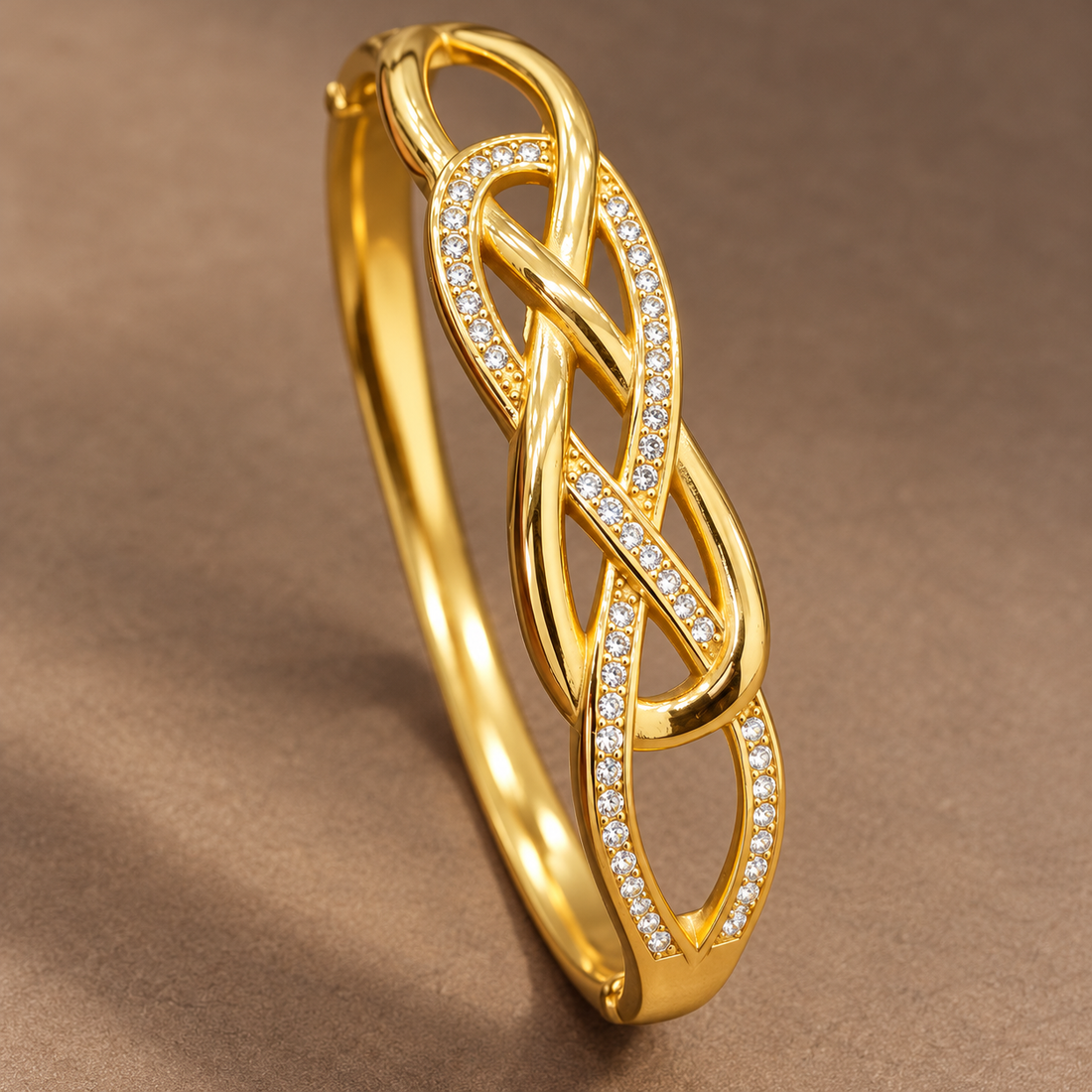 Gold-tone 304 stainless steel bangle bracelet with a woven infinity-knot top set with clear crystals and a hinged clasp, lying flat on a brown surface