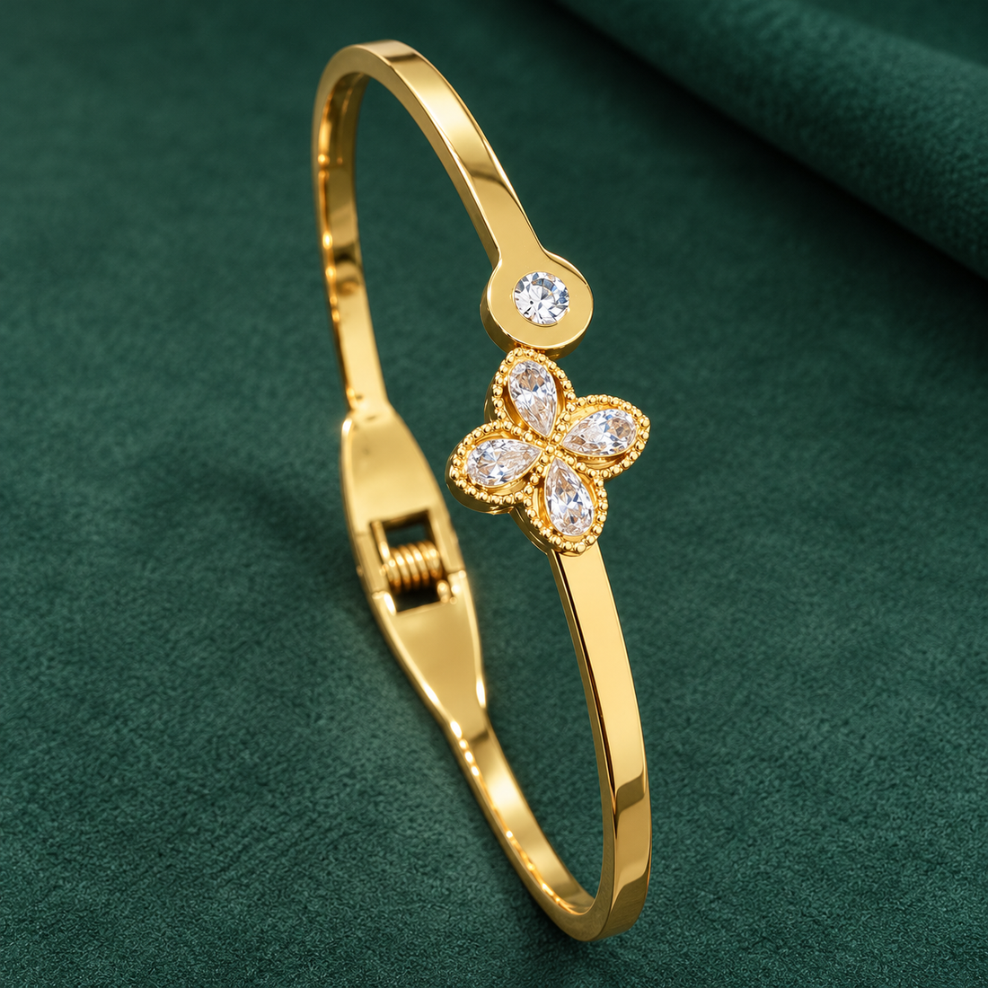 Gold-tone 304 stainless steel hinged bangle with a four-petal flower of clear pear-cut stones and a round stone on the clasp end, on a green cloth