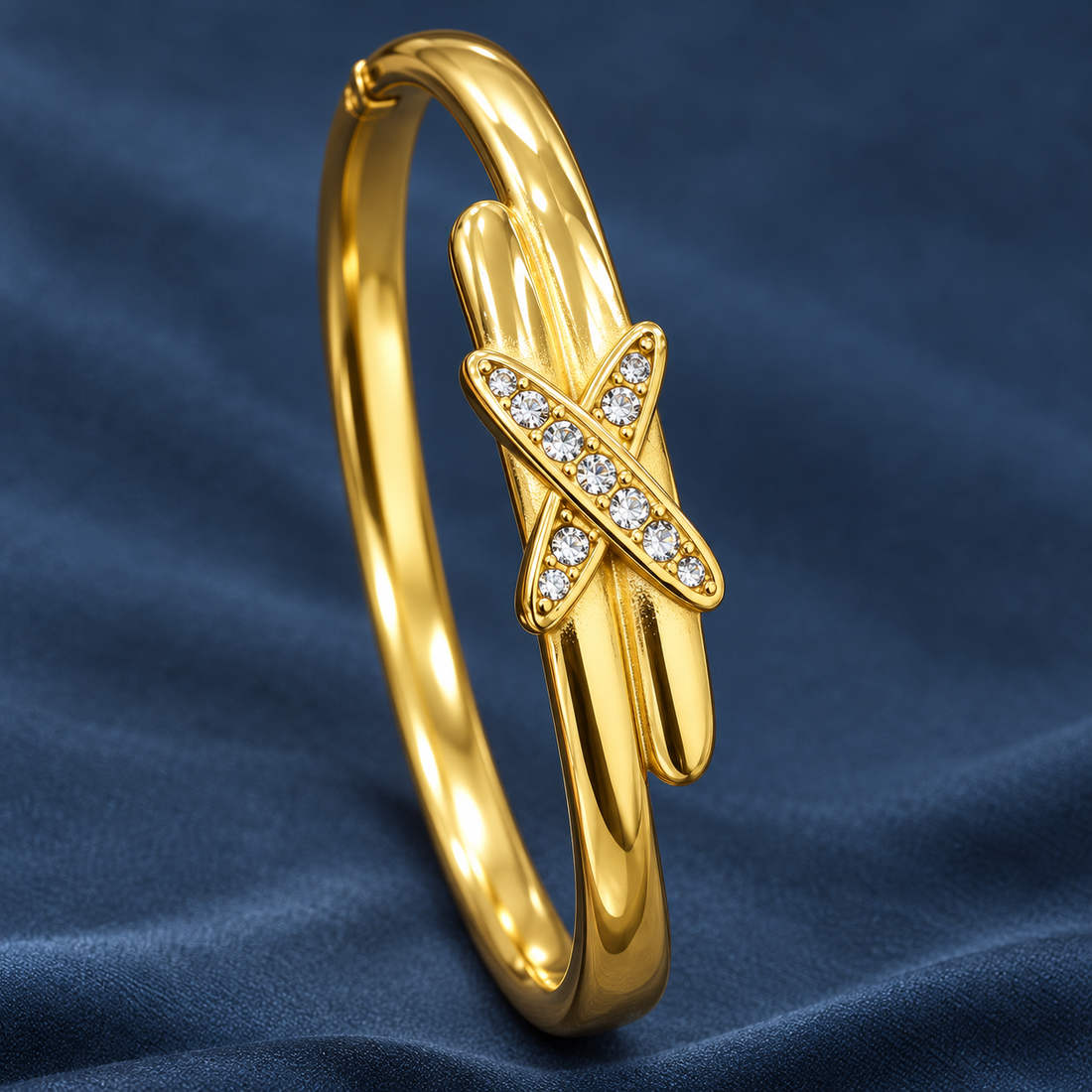 Gold-tone 304 stainless steel bangle bracelet with a crossed-band front set with clear crystals, shown on a dark blue fabric