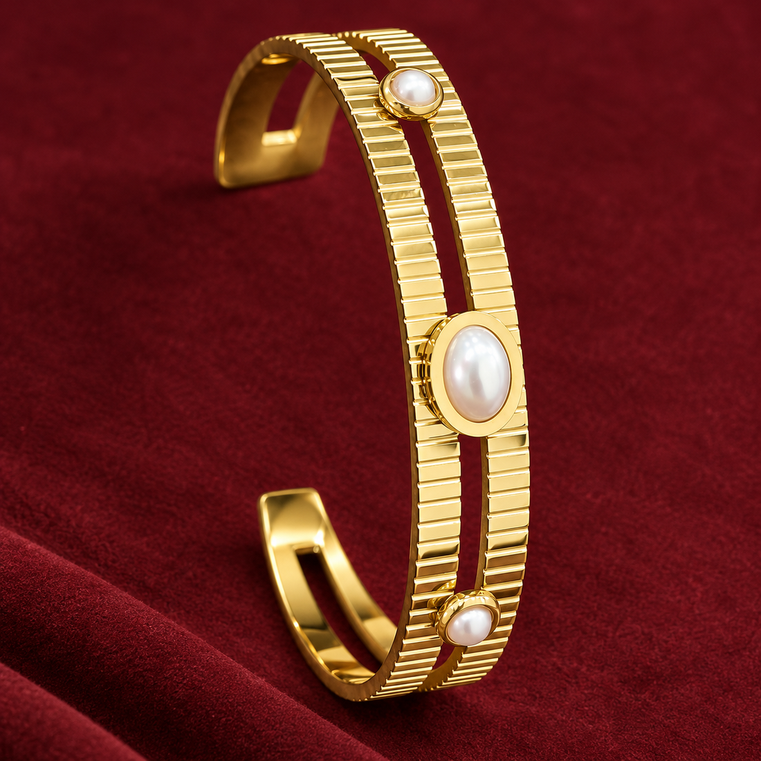 Gold-tone 304 stainless steel cuff bracelet with a ribbed open band and three white pearl-style stones set in bezels, on a dark red velvet backdrop