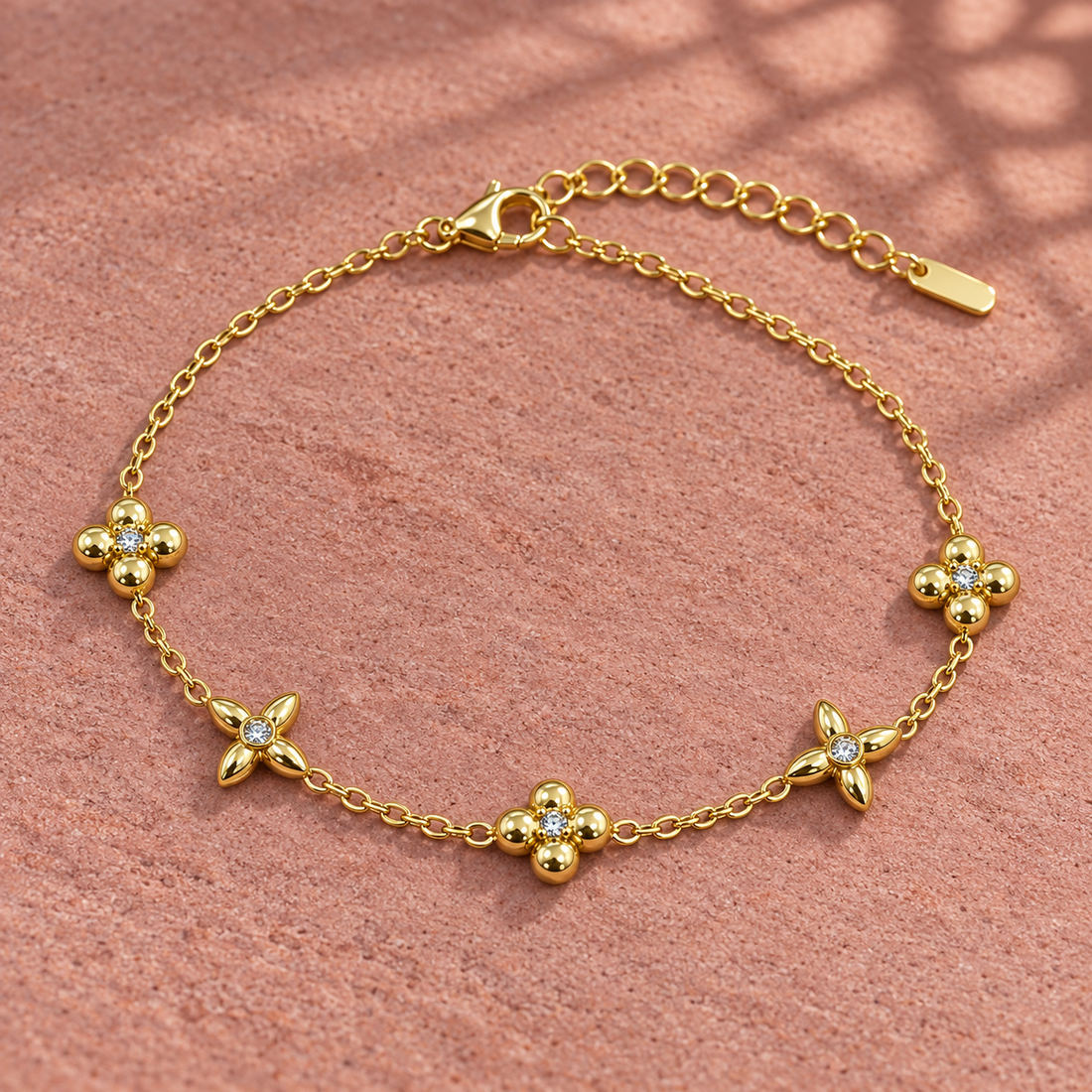 Gold-tone 316L stainless steel chain bracelet with five flower motifs set with small crystals, lying flat on a pink surface