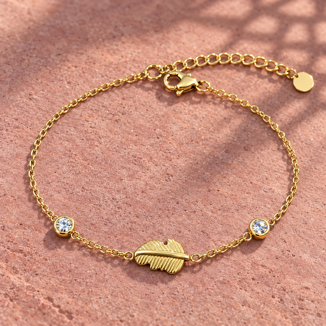 Gold-tone 316L stainless steel chain bracelet with a textured feather charm and two round clear stones, laid flat on a terracotta surface