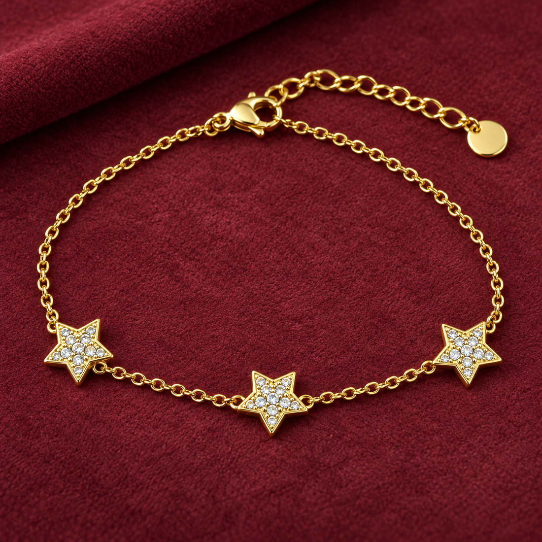 Gold-tone 316L stainless steel chain bracelet with three star charms set with clear stones and a lobster clasp, lying flat on red fabric