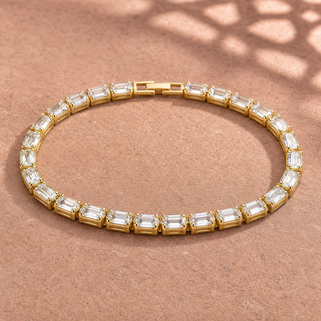 Gold-tone brass tennis bracelet with a row of emerald-cut clear stones in claw settings and a box clasp, lying flat on a sandstone surface