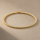 Gold-tone brass tennis bracelet with a row of clear stones in claw settings and a box clasp, laid flat on a beige surface