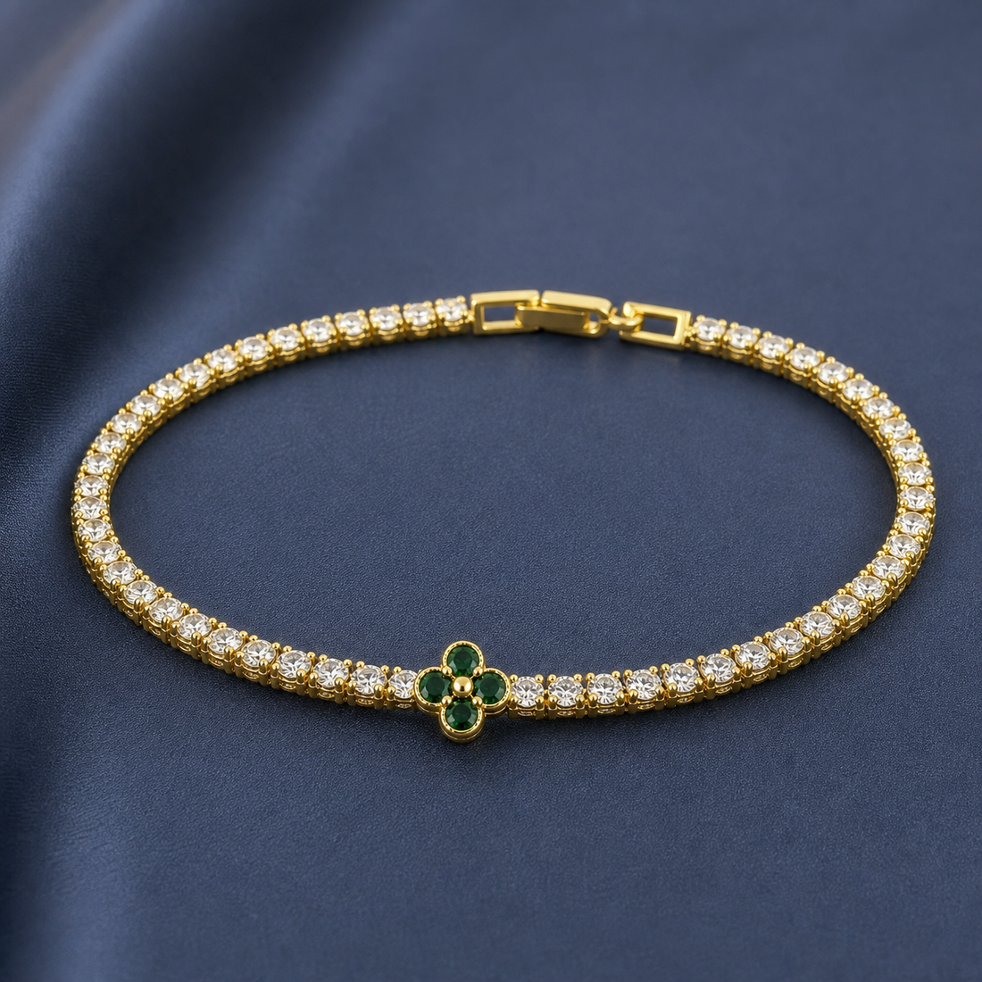 Gold-tone brass tennis bracelet with clear stones and a four-petal clover set with green stones, lying flat on a navy background