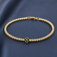 Gold-tone brass tennis bracelet with clear stones and a four-petal clover set with green stones, lying flat on a navy background