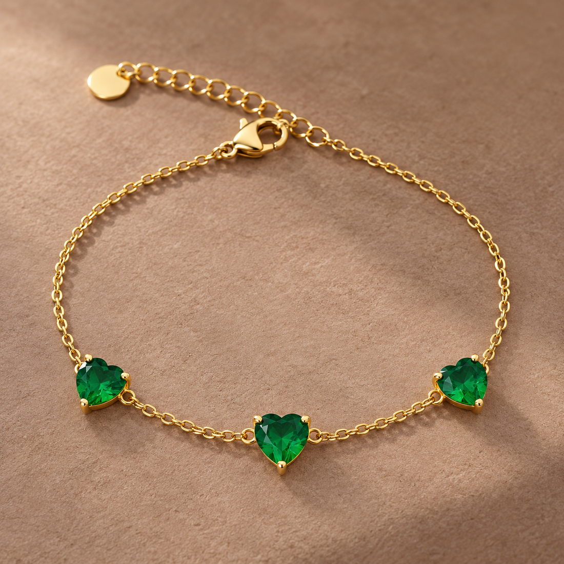 Gold-tone 316L stainless steel chain bracelet with three green heart-cut stones in claw settings and a lobster clasp, laid flat on a tan surface