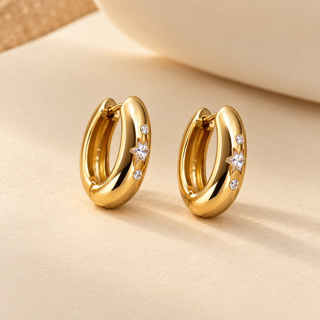 Gold-tone 316L stainless steel huggie hoop earrings with a star-shaped and round clear stone set on each, shown as a pair on a beige surface