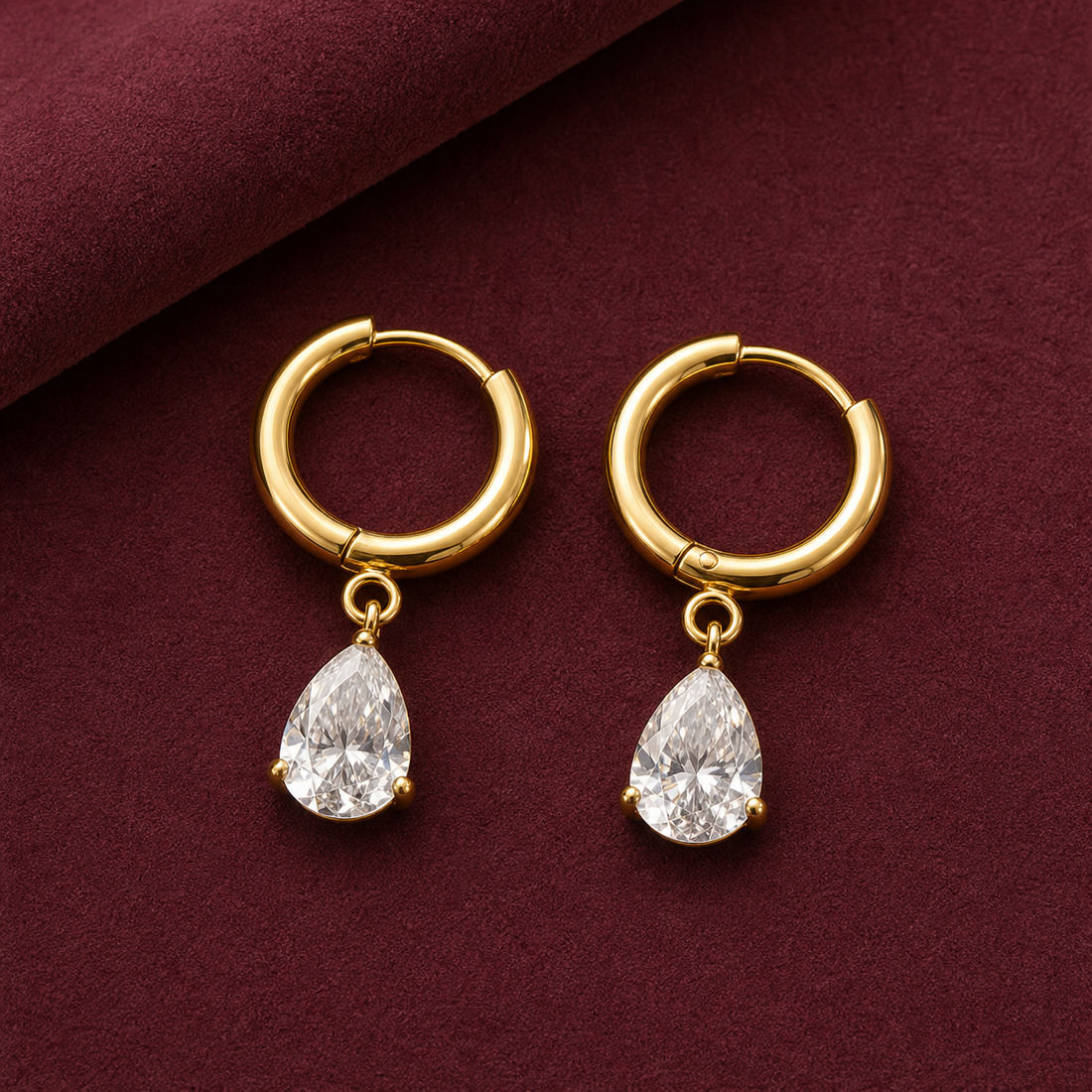 Gold-tone 316L hoop earrings with a pear-cut clear crystal drop on a pair, shown flat on a burgundy surface, a second angle