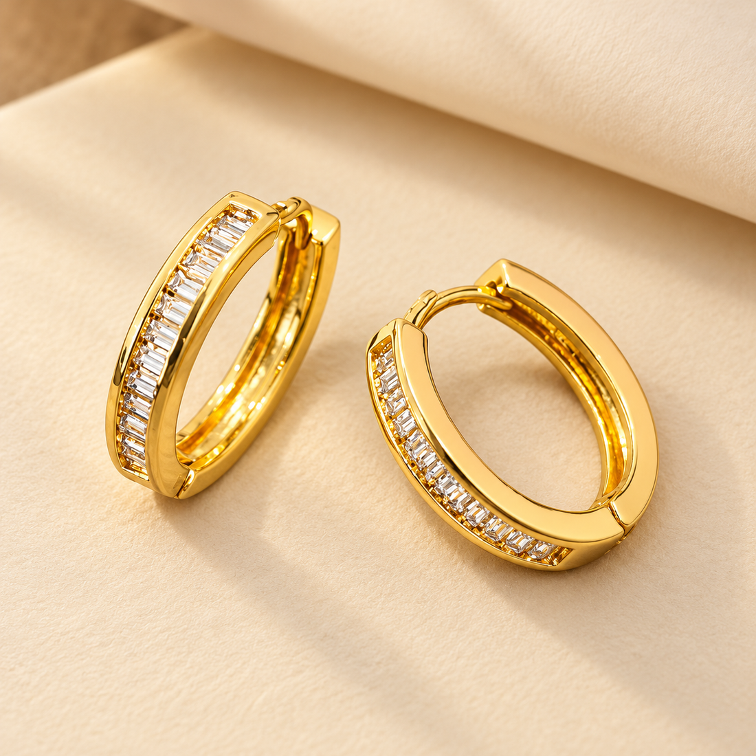 Gold-tone 316L stainless steel huggie hoop earrings with a row of baguette-cut clear crystals along the outer edge, a pair shown on a cream surface