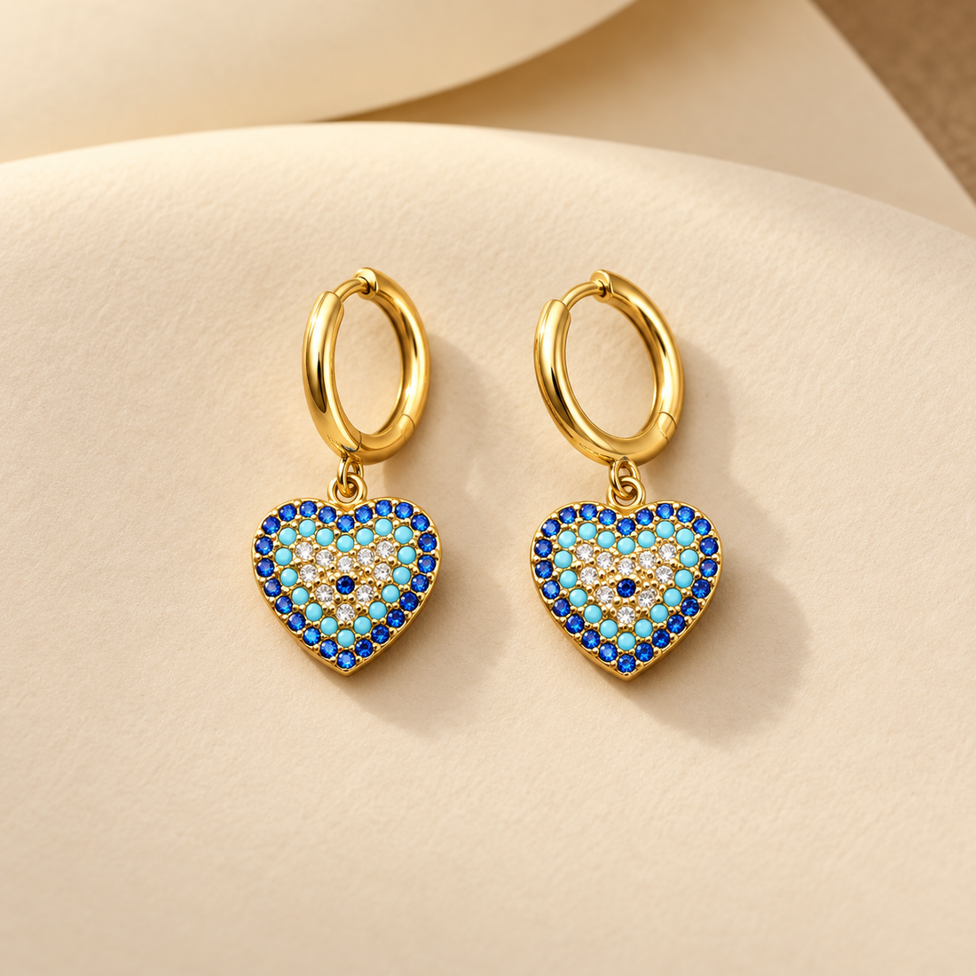 Gold-tone 316L stainless steel huggie earrings with heart drops set with blue, turquoise and white stones around a blue centre, shown as a pair