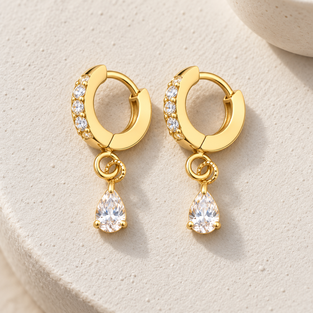 Gold-tone 316L stainless steel huggie hoop earrings with a row of clear stones on the top half and a pear-cut drop stone, a pair on a stone surface