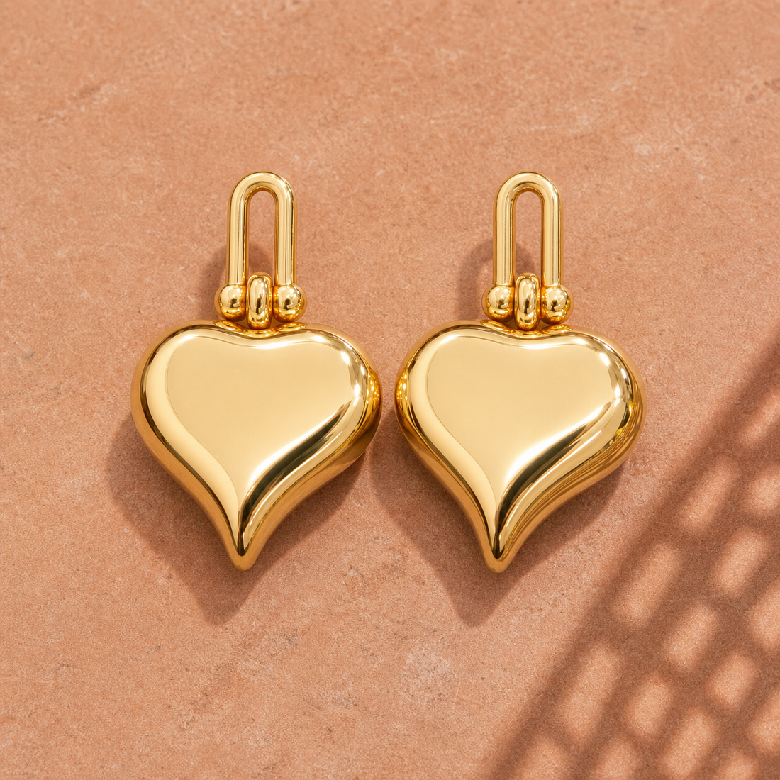 Gold-tone 304 stainless steel earrings with puffed heart drops hanging from a looped link top, a pair shown flat on a sandstone surface