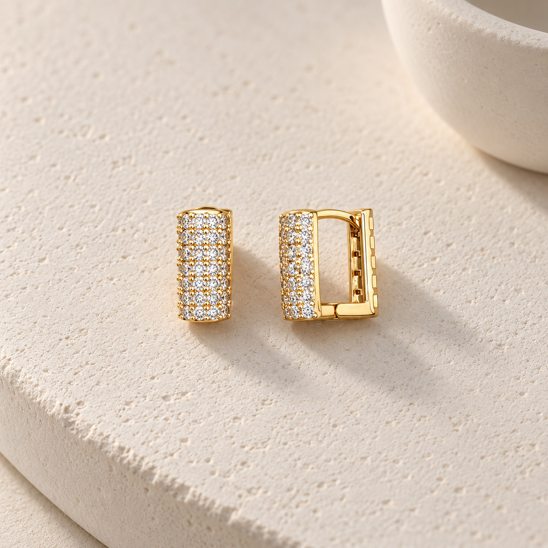Gold-tone 316L stainless steel huggie earrings with rows of clear crystals across a square-profile band, shown as a pair on a stone surface