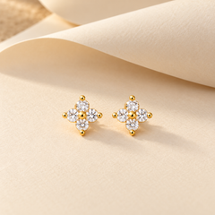 Gold-tone 316L stainless steel stud earrings with a four-petal cluster of clear crystals and gold ball accents, a pair shown on a cream background