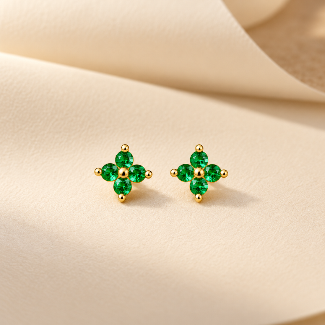 Gold-tone 316L stainless steel stud earrings with four green stones in a cross cluster and a gold-tone centre bead, shown as a pair on a cream fabric background