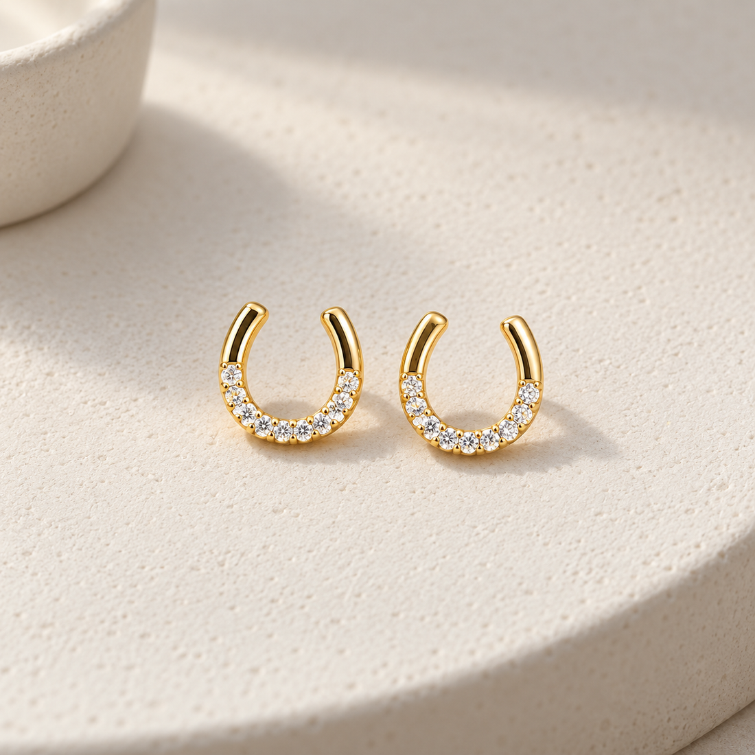 Gold-tone 304 stainless steel pair of open horseshoe-shaped cuff earrings set with clear crystals along the lower curve, shown on a cream stone surface
