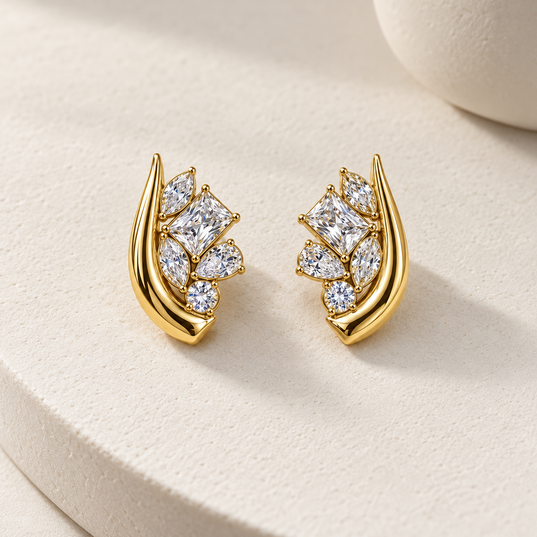 Gold-tone 316L stainless steel stud earrings pair with a curved swirl and clustered clear stones in marquise, princess, pear and round cuts, shown as a pair