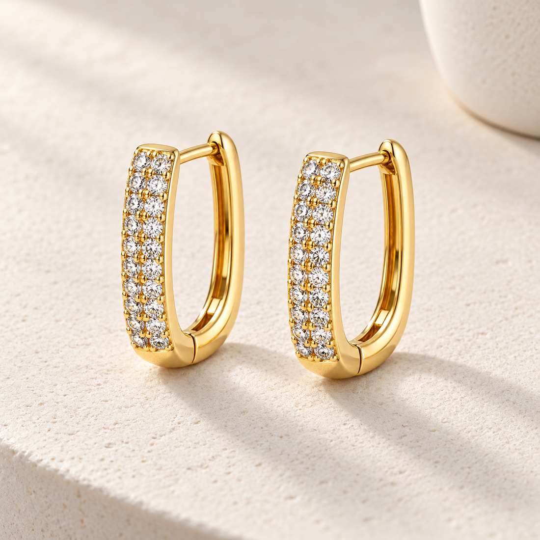 Gold-tone 316L stainless steel pair of U-shaped huggie hoop earrings with two rows of clear crystals along the front, shown on a cream surface