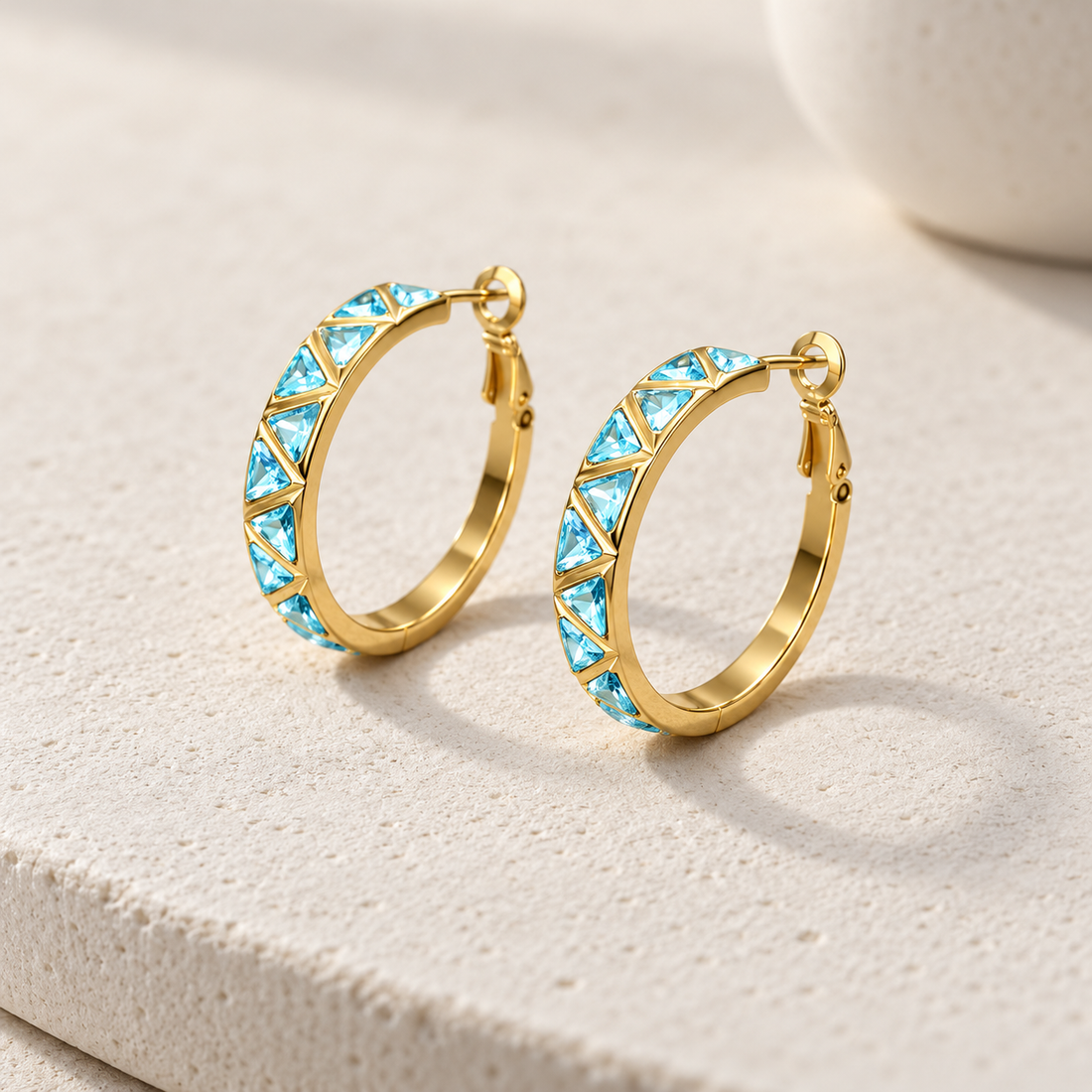 Gold-tone 316L stainless steel hoop earrings with a row of aqua triangular stones set along the front, a pair shown on a light stone surface