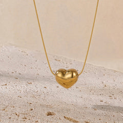 A gold heart-shaped pendant necklace hanging from a delicate gold chain against a warm beige textured background.