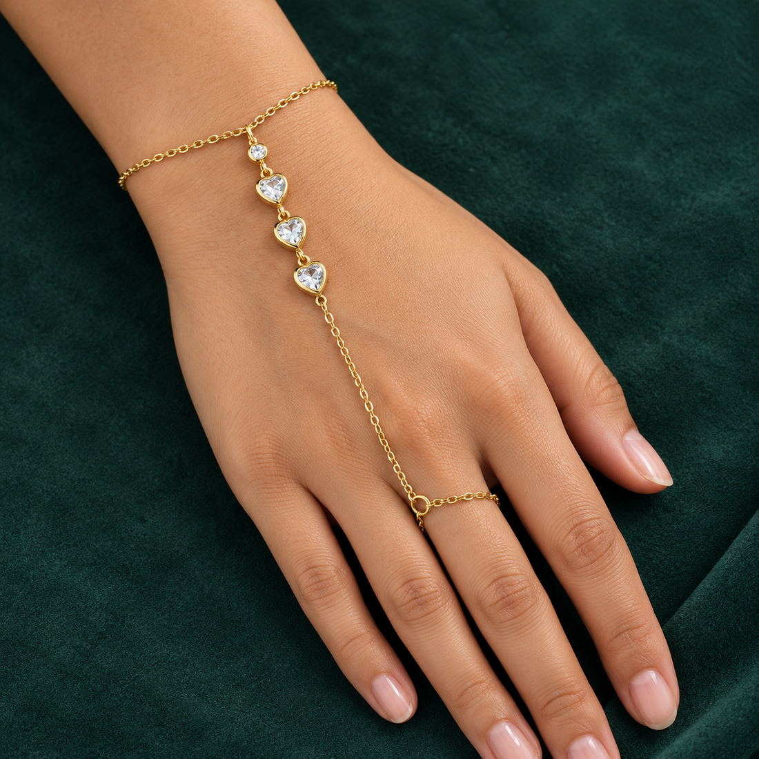 Gold-tone 304 stainless steel hand chain bracelet with three heart-cut clear stones and a small round stone, joined to a finger ring, worn on a hand
