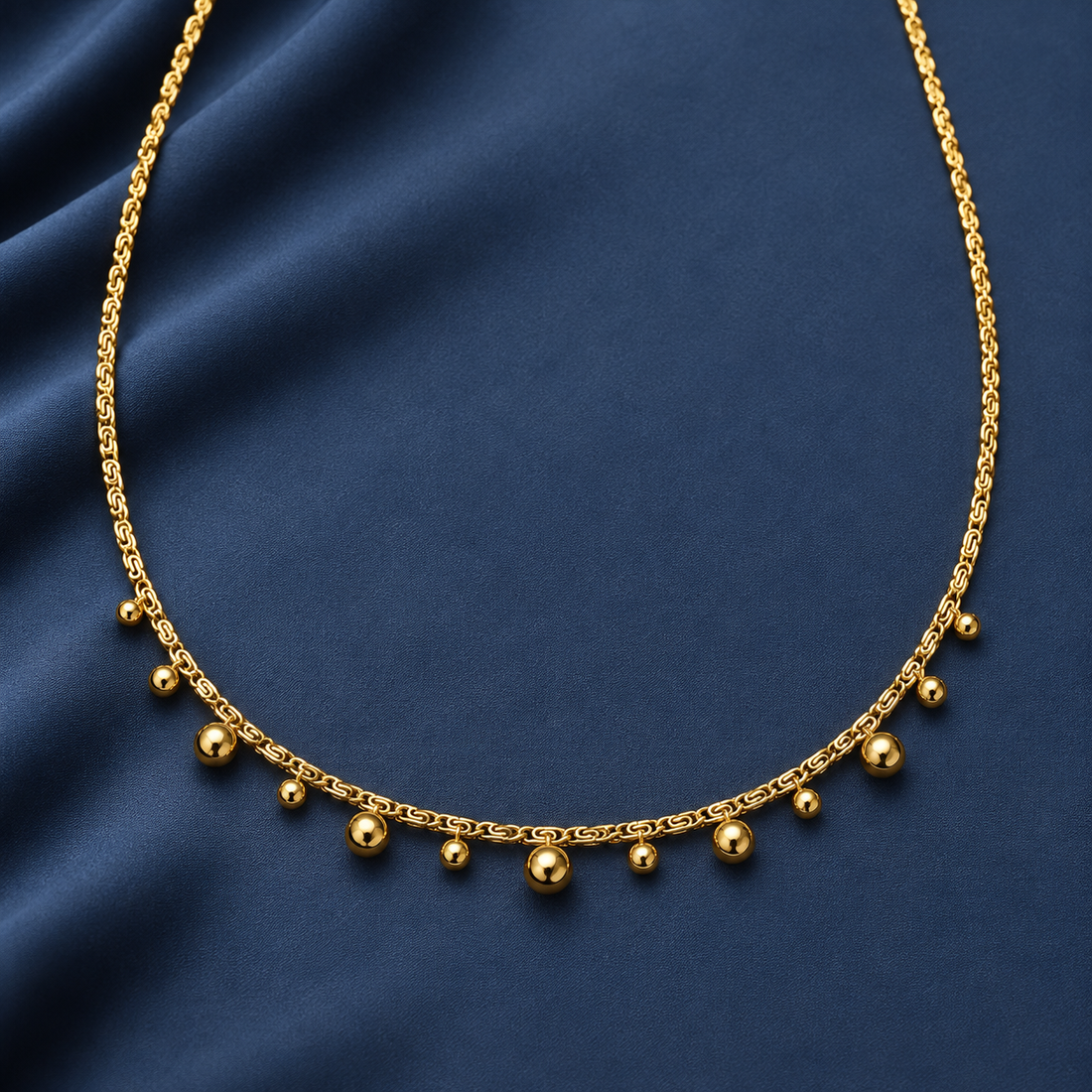 Gold-tone 316L stainless steel necklace with a fine link chain and graduated ball drops along the front, laid flat on a dark blue cloth