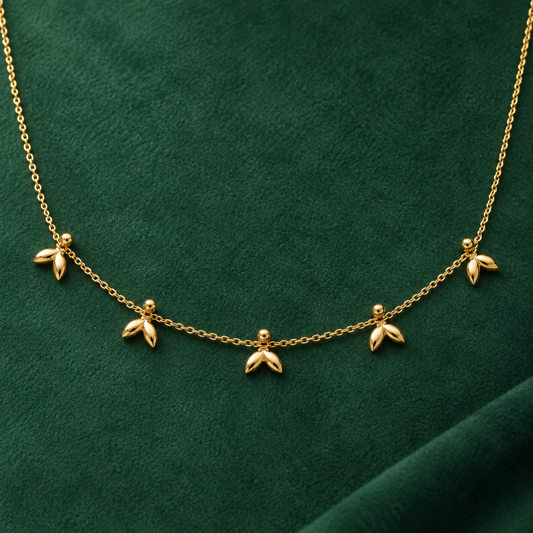 Gold-tone 316L stainless steel necklace with a fine cable chain and five small leaf-cluster drops, shown laid flat on green velvet