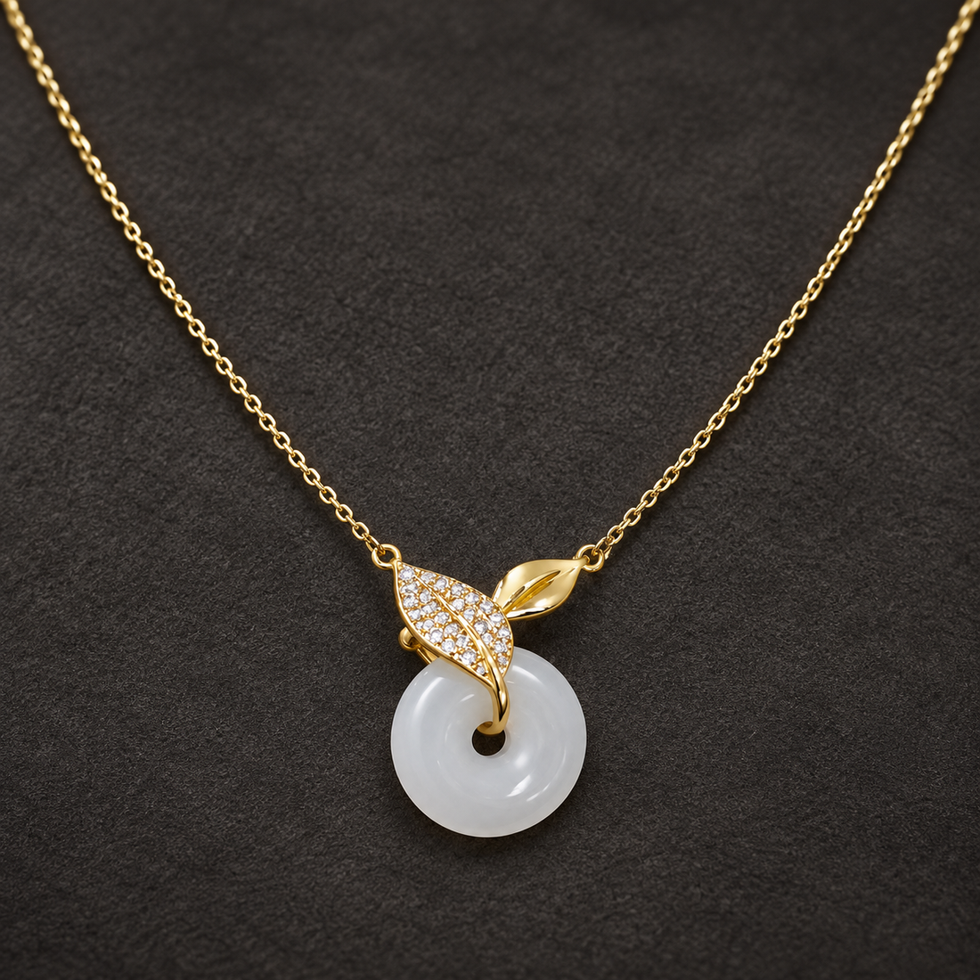 Gold-tone 316L stainless steel necklace with a white jade-look donut pendant and a leaf-shaped crystal-set bail on a fine cable chain, laid flat on dark fabric