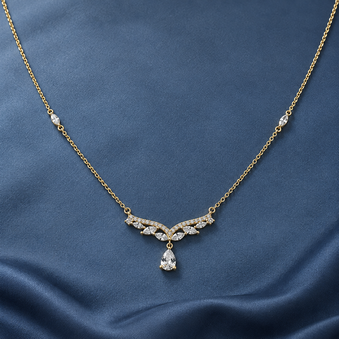 Gold-tone 316L stainless steel necklace with a V-shaped row of clear stones and a pear-cut drop pendant, laid flat on blue fabric