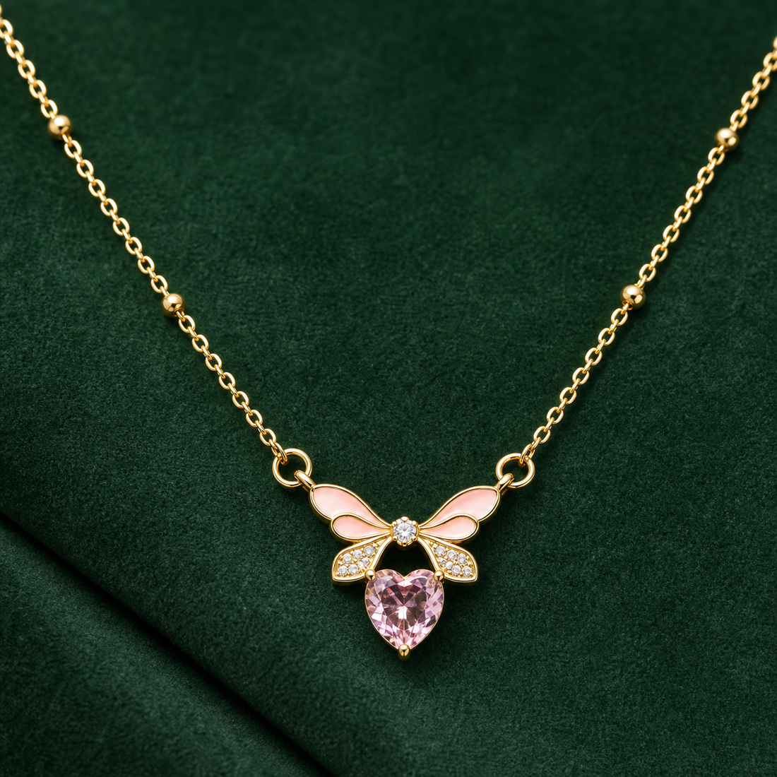 Gold-tone 304 stainless steel necklace with a pink enamel bow and a pink heart-cut stone, shown lying on green fabric