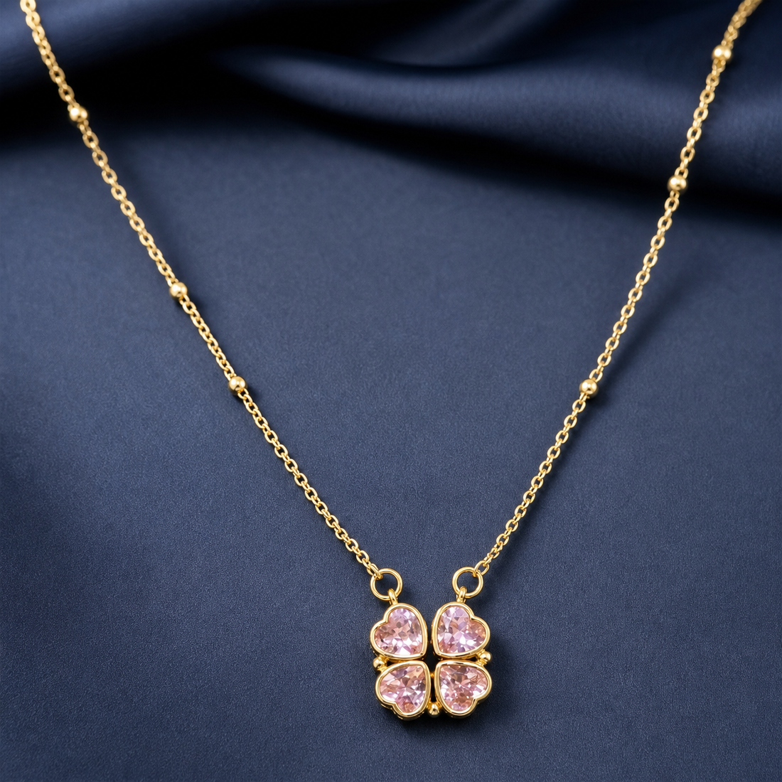 Gold-tone 304 stainless steel necklace with a four-leaf clover pendant of pink heart-cut stones on a satellite bead chain, shown laid on navy fabric