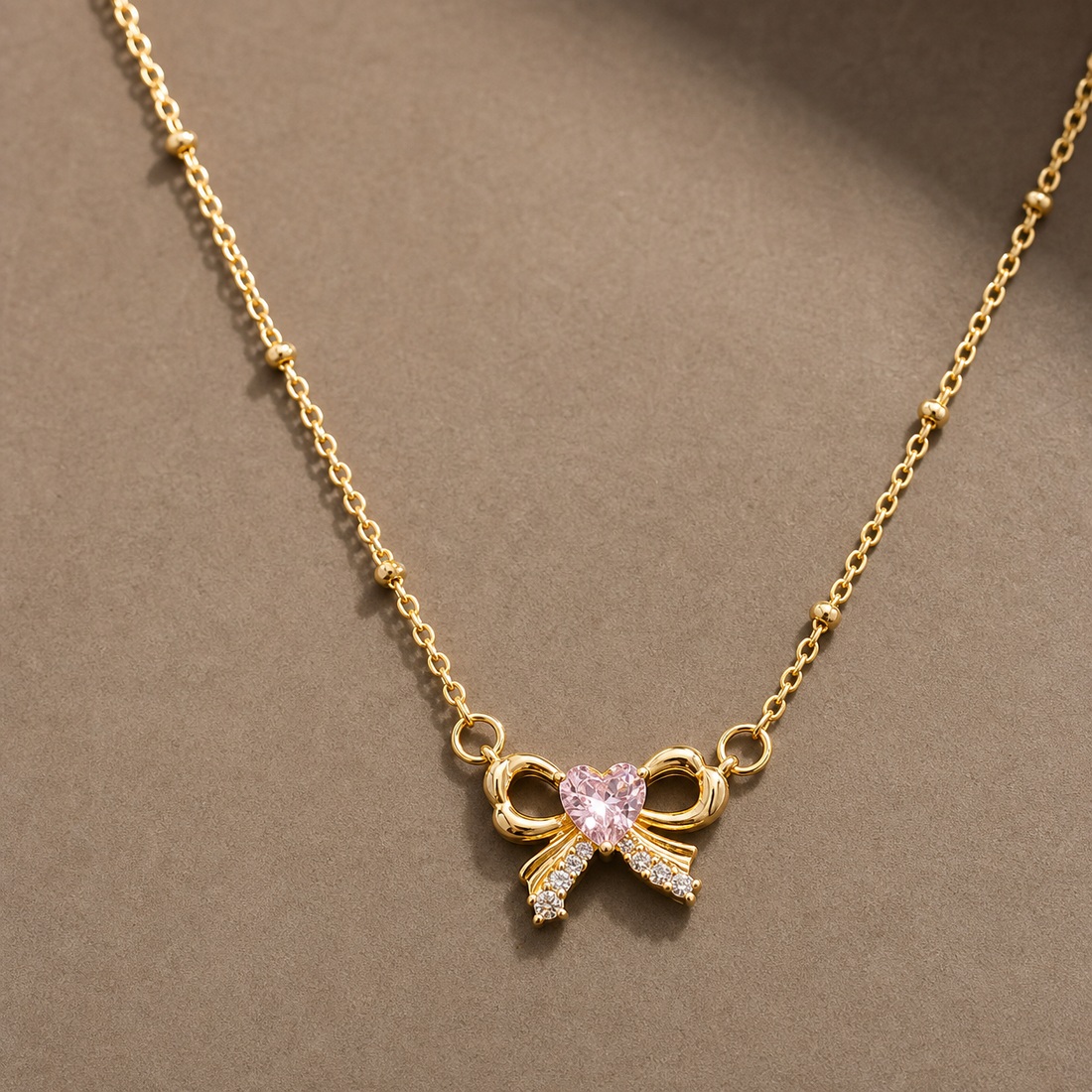 Gold-tone 304 stainless steel necklace with a bow pendant set with a pink heart stone and clear stones along one loop, on a bead-accent chain