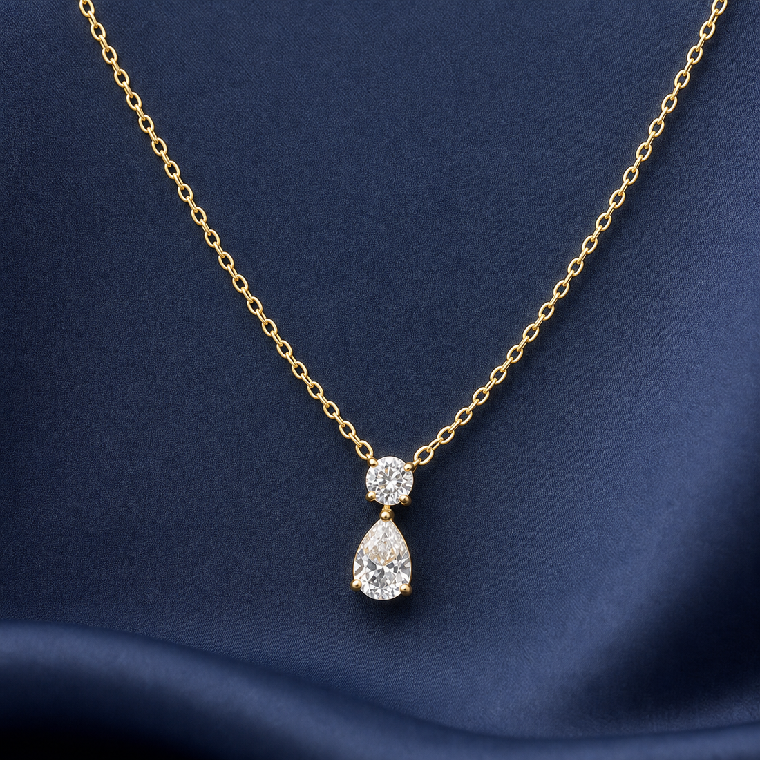 Gold-tone 316L pendant necklace with a round and a pear-cut clear stone drop on a fine cable chain, displayed on navy fabric