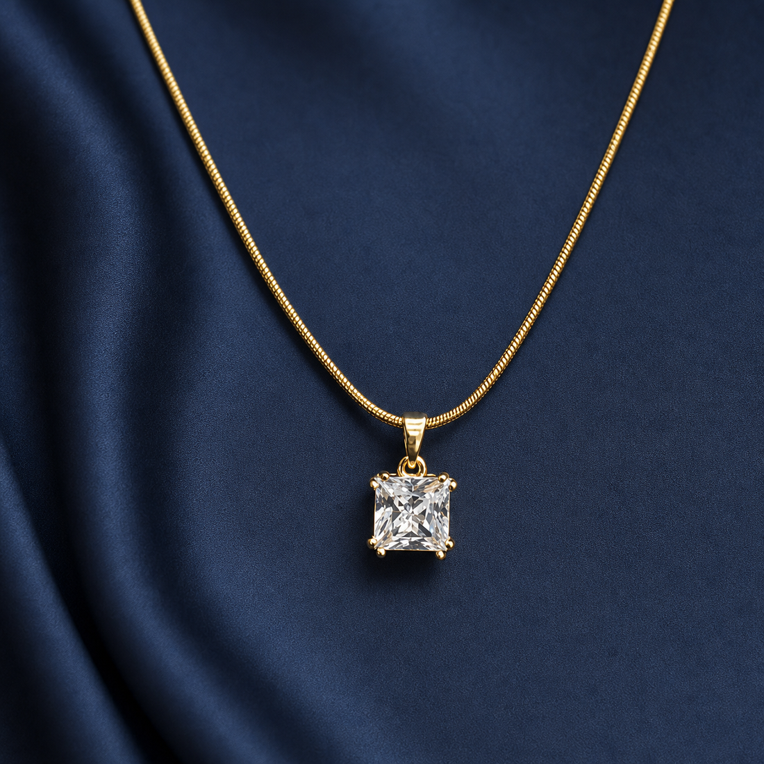 Gold-tone 316L stainless steel snake chain necklace with a square crystal pendant in a four-prong setting, worn on a navy fabric backdrop