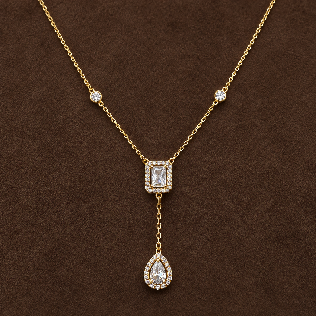 Gold-tone 316L stainless steel necklace with a clear stone-set rectangle and teardrop pendant on a Y-drop chain, laid flat on a brown surface