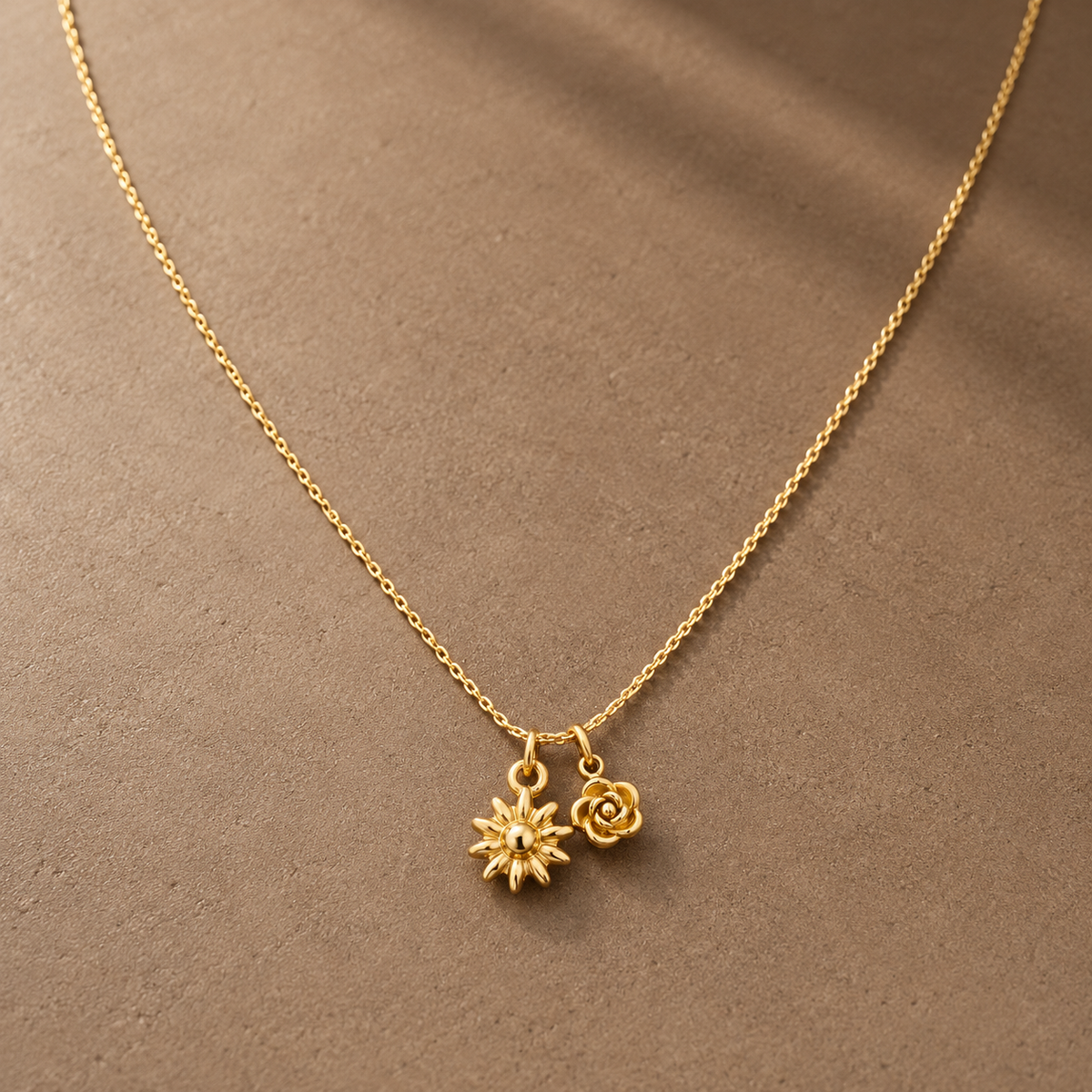 Gold-tone 316L stainless steel necklace with a fine cable chain and two small flower charms, a daisy and a rose, laid flat on a fabric surface