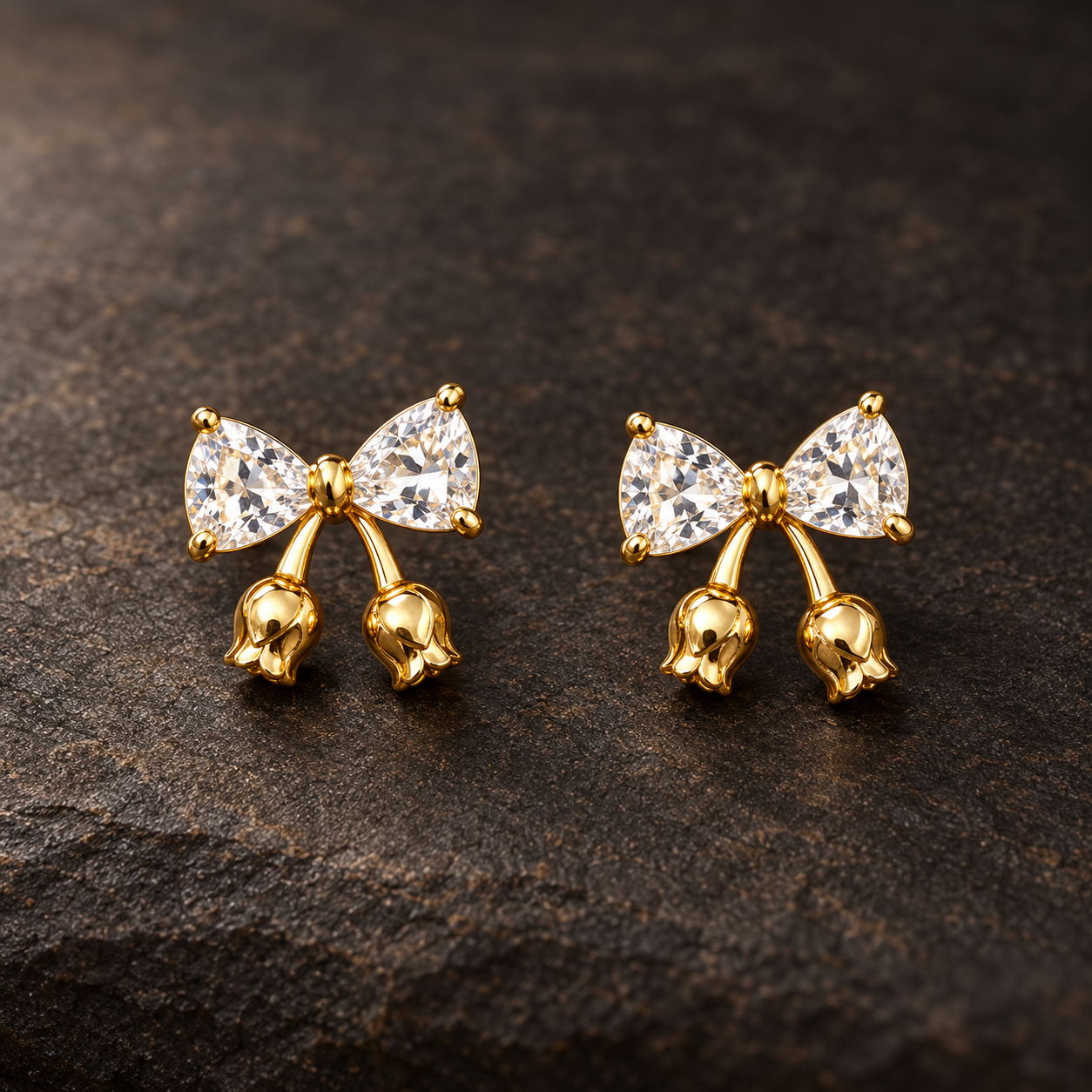 Gold-tone 304 stainless steel earrings with a bow of clear trillion-cut stones and small gold tulip-bud drops, a pair on a dark stone surface