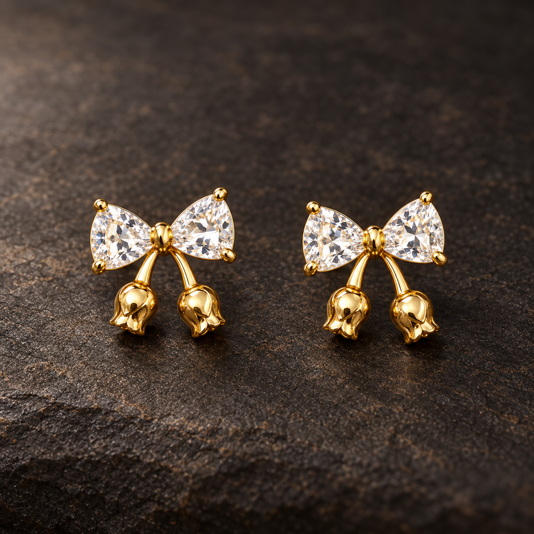 Gold-tone 304 stainless steel earrings with a bow of clear trillion-cut stones and small gold tulip-bud drops, a pair on a dark stone surface