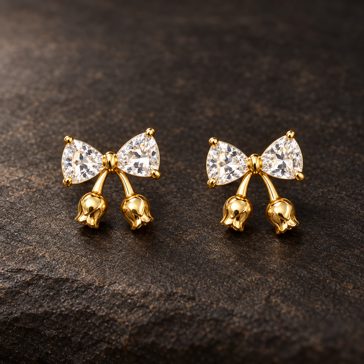 Gold-tone 304 stainless steel earrings with a bow of clear trillion-cut stones and small gold tulip-bud drops, a pair on a dark stone surface