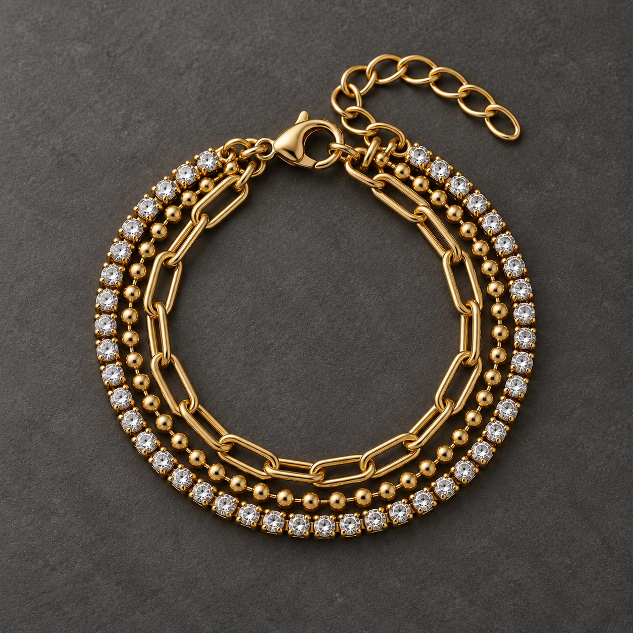 Gold-tone 316L stainless steel layered bracelet set with a crystal tennis band, a ball chain and a paperclip chain, laid flat on a dark surface