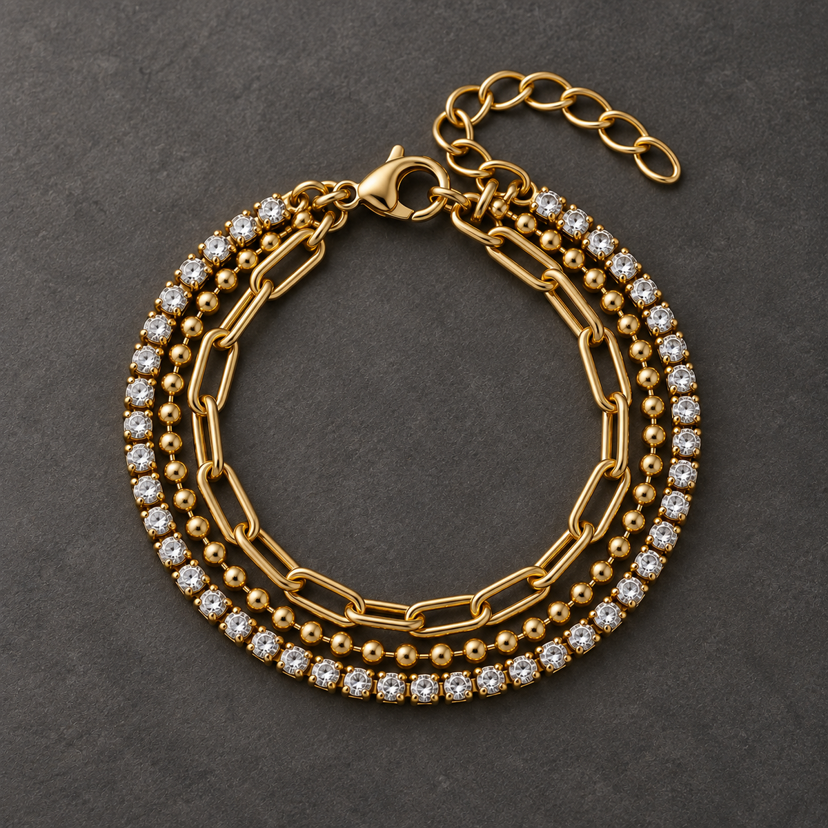 Gold-tone 316L stainless steel layered bracelet set with a crystal tennis band, a ball chain and a paperclip chain, laid flat on a dark surface