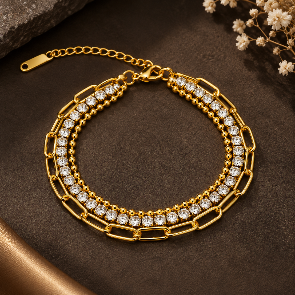 Gold-tone 304 stainless steel layered bracelet with a paperclip chain, a bead chain and a row of clear crystal stones, lying flat on a dark surface