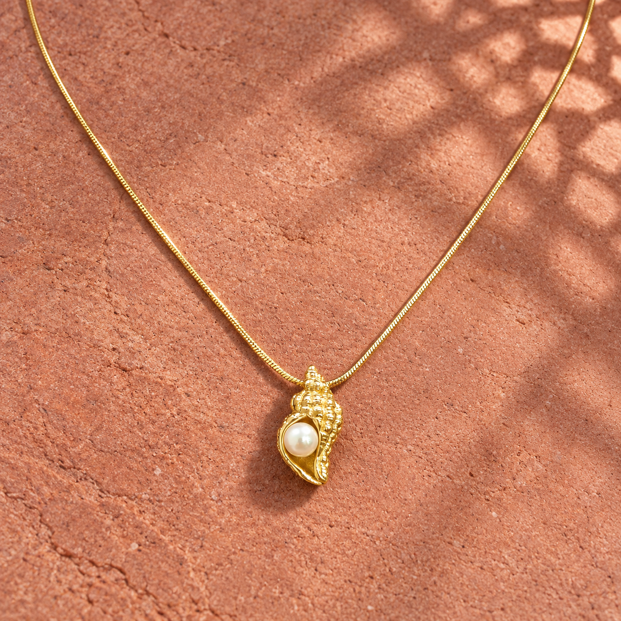 Gold-tone 316L stainless steel snake chain necklace with a textured conch shell pendant set with a cream pearl, laid on a terracotta surface