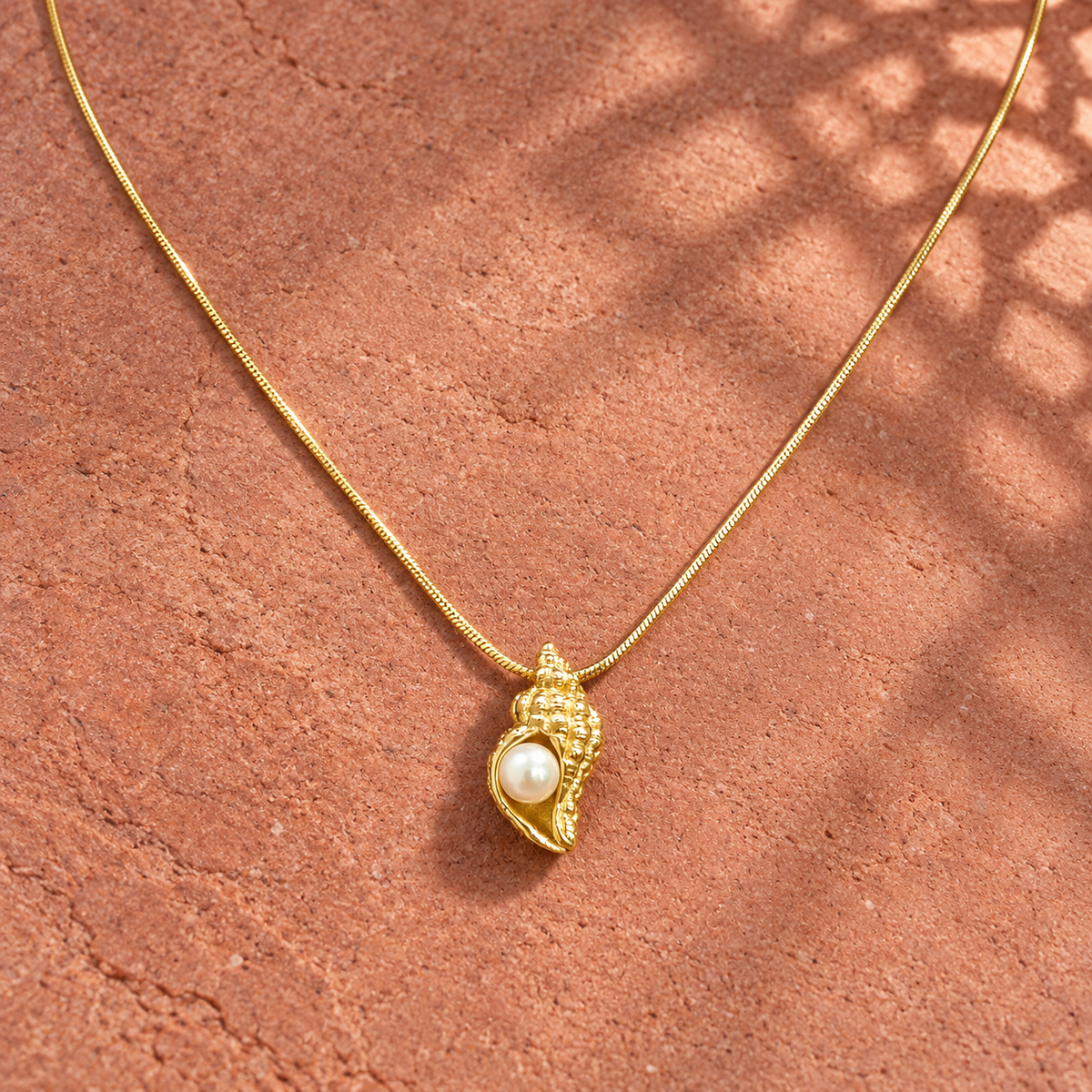 Gold-tone 316L stainless steel snake chain necklace with a textured conch shell pendant set with a cream pearl, laid on a terracotta surface