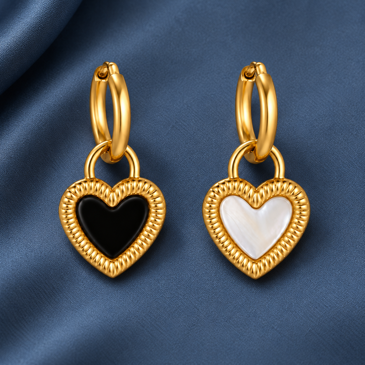 Gold-tone 316L stainless steel hoop earrings with a ridged heart charm, one black and one white mother-of-pearl-look inlay, a pair on blue fabric