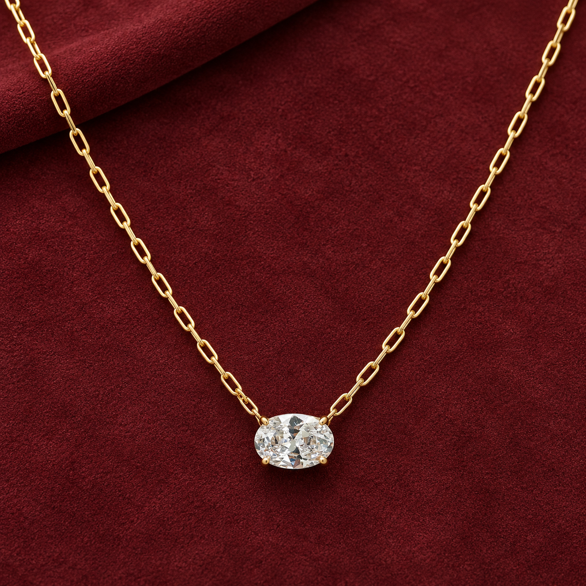 Gold-tone 316L stainless steel necklace with a paperclip chain and an oval clear crystal pendant in a four-prong setting, laid on a dark red cloth