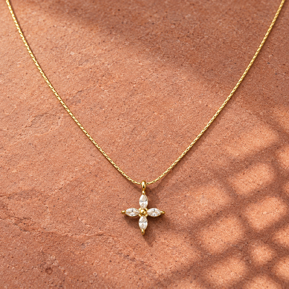 Gold-tone 316L stainless steel necklace with a fine chain and a cross pendant set with four marquise stones, lying on a stone surface