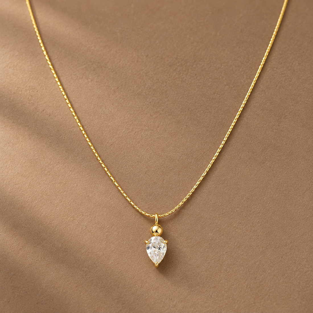 Gold-tone 316L stainless steel necklace with a fine textured chain and a pear-cut clear crystal pendant on a small ball bail, worn at the neckline